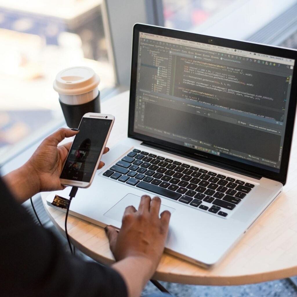 Developer working remotely, coding on a laptop with phone in hand, showcasing modern work culture.